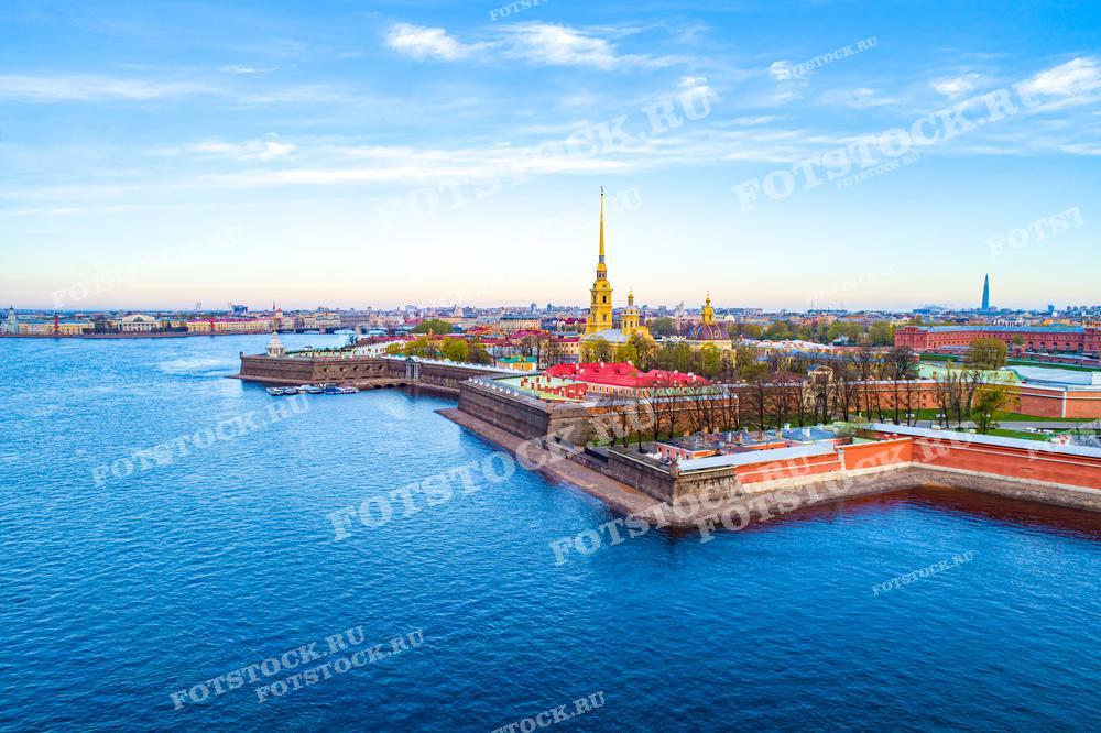 Фотография демонстрирует вид на Петропавловскую крепость в Санкт-Петербурге, снятый с воздуха. Крепость расположена на берегу реки Невы и является одним из ключевых исторических памятников города. Видны высокий шпиль Петропавловского собора и окружающие его здания, а также река и городской пейзаж на заднем плане. Небо ясное, что делает изображение ярким и насыщенным. Это фото может использоваться в культурных и туристических публикациях, образовательных материалах, а также в качестве элемента урбанистического дизайна и городской планировки.